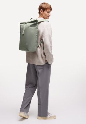 Giovane che indossa pantaloni grigi, maglione beige, sneakers bianche, e porta uno zaino grande a rotolo verde chiaro su una spalla.