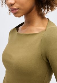 Gros plan sur une femme portant un haut à manches longues vert olive avec un large décolleté carré et des cheveux bouclés tirés en arrière.