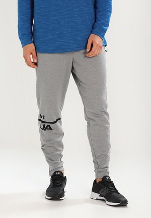 Uomo che indossa pantaloni della tuta grigi Under Armour e scarpe da corsa nere, in piedi con le mani rilassate lungo i fianchi.