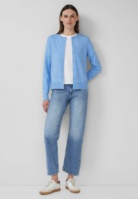 Lichtblauwe cardigan met ronde hals en knoopsluiting, gedragen over een wit T-shirt. Gepaard met lichtblauwe rechte jeans en witte sneakers.