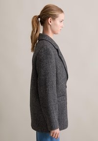 Szary żakiet w kratę houndstooth z dużymi klapami, pojedynczym guzikiem i kieszeniami bocznymi. Teksturowana tkanina o luźnym kroju, noszona na niebieskich dżinsach.