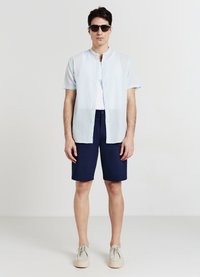 Chemise bleu clair à manches courtes avec un col mao, t-shirt blanc en dessous, short bleu marine, et baskets beiges. Lunettes de soleil noires en accent.