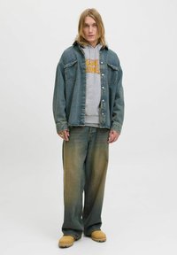 Veste en denim à la coupe décontractée, délavée bleu clair, avec des poches à bouton, portée par-dessus un sweat à capuche gris avec impression graphique, associée à un jean à jambe large.