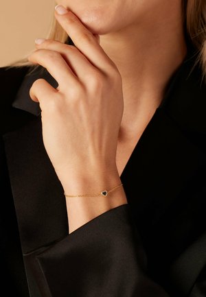 Bracelet en chaîne dorée avec un petit charme en forme de cœur noir, porté sur une main contre un fond de tissu sombre. Design élégant et délicat.