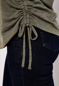 Top verde texturizado con lados fruncidos, que presenta un detalle de cordón. Combinado con jeans de mezclilla oscuros que muestran un diseño en el bolsillo trasero.