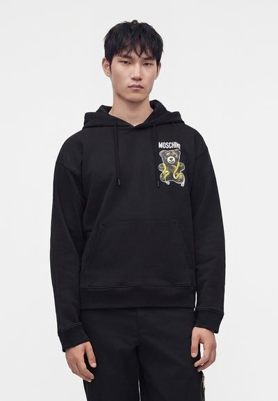 Νεαρός άνδρας που φοράει μαύρο φούτερ Moschino με ένα κίτρινο και μαύρο σχέδιο αρκουδάκι στο στήθος, στέκεται μπροστά σε λευκό φόντο.