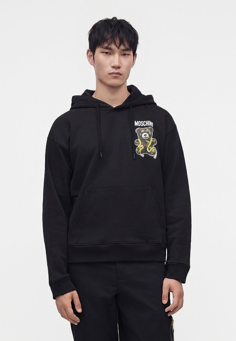 Νεαρός άνδρας που φοράει μαύρο φούτερ Moschino με ένα κίτρινο και μαύρο σχέδιο αρκουδάκι στο στήθος, στέκεται μπροστά σε λευκό φόντο.
