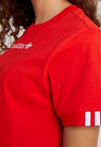 Röd bomull t-shirt med rund halsringning, med vitt Adidas-logotyp på bröstet och tre vita ränder på ärmarna.
