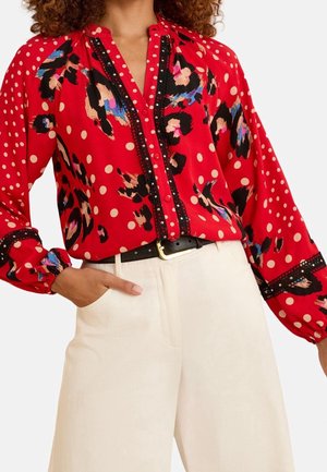 Femme portant un chemisier rouge à manches longues avec des motifs à pois et floraux abstraits, associé à un pantalon blanc et une ceinture noire.