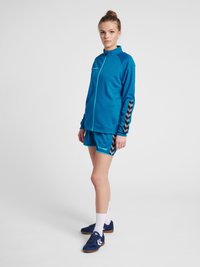 Blauwe sportjacket met een volle rits en chevronpatroon op de mouwen, gecombineerd met bijpassende shorts. Draag het met marine blauwe schoenen en witte sokken.