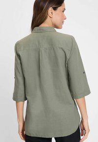 Olive green, lightweight shirt with three-quarter sleeves, rounded hem, and back pleat detail. Features a classic collar and buttoned cuffs.