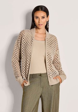 Frau trägt eine beige offene Strickjacke über einem beigen Oberteil und olivgrünen Fischgrät-Hosen und steht mit den Händen in den Taschen.