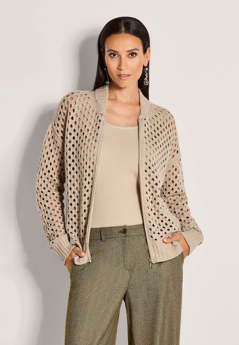 Vrouw in een beige opengehaakte cardigan over een beige top en olijfgroene herringbone broek, staande met haar handen in de zakken.