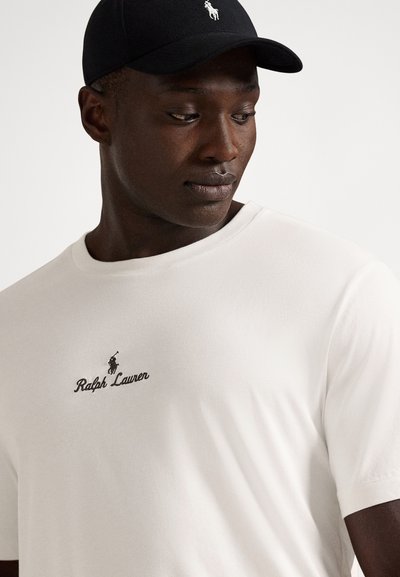 Hvit bomulls-T-skjorte med rund hals, med sort brodert "Ralph Lauren"-logo og et lite svart hestemerke. Svart caps på hodet.