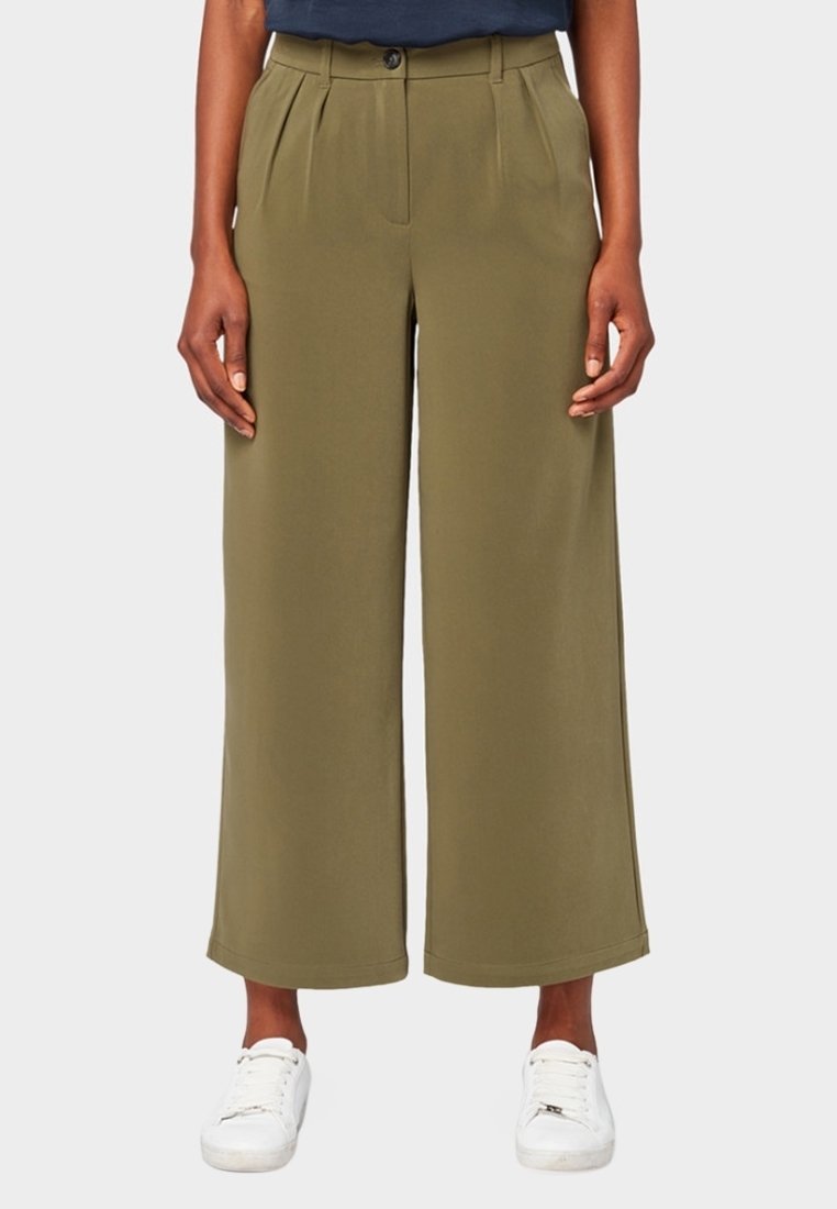 Personne portant un pantalon large raccourci vert olive avec des plis à l'avant et des baskets blanches, debout devant un fond uni.