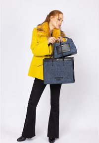 Gul kappa med pälskrage, svarta byxor, håller en blå texturerad väska och en mindre grå väska med svarta detaljer. Svarta skor.