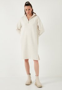 Helles beiges Oversized-Kleid mit einem Halb-Reißverschluss-Kragen; lange Ärmel und seitliche Schlitze. Das Model kombinierte es mit cremefarbenen Ankle Boots. Weicher, texturierter Stoff.