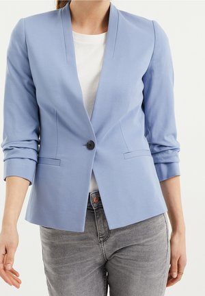 Vrouw draagt een lichtblauwe blazer met één knoop en geplooide mouwen, wit topje en grijze spijkerbroek tegen een effen achtergrond.