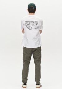 T-shirt blanc à manches courtes avec un graphisme noir représentant des papiers pliés au dos. Un pantalon gris texturé et des chaussures noir et blanc complètent le look.