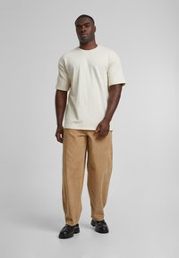 Crème katoenen t-shirt met korte mouwen, gecombineerd met beige corduroy broeken en zwarte chunky schoenen. Model staat op een effen achtergrond.