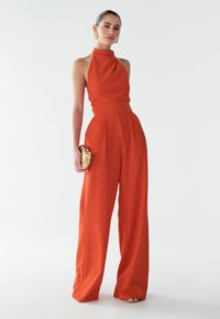 Červený overal s halter výstřihem, širokými nohavicemi, texturovanou látkou, staženým pasem a složitou zlatou kabelkou. Vyznačuje se minimalistickým designem a výraznou barvou.
