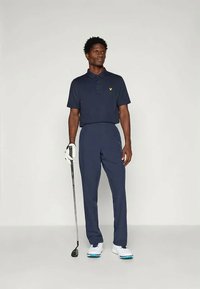 Polo de golfe azul-marinho com logótipo amarelo, combinado com calças a condizer. O modelo segura um taco de golfe, usando sapatos brancos com detalhes em azul.