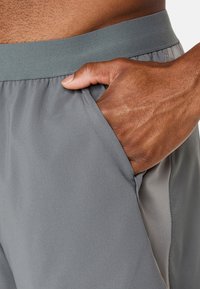 Grå atletiske shorts med en glat tekstur og elastisk talje, der har en sidelomme og lette mesh-detaljer langs sømmen.