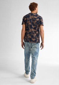 Camisa navy con patrón floral en beige, de manga corta, combinada con jeans en azul claro y zapatillas blancas, vista desde atrás.