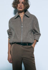 Ruskea vetoketjullinen takki, jossa on kaulus ja kuvioitu kangas, yhdistettynä ruskeisiin housuihin ja mustaan vyöhön. Asussa on rento hiha.