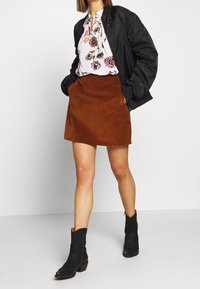 Gonna in velluto a coste marrone con tasche laterali, abbinata a una blusa floreale bianca e a una giacca bomber nera. Stivaletti neri alla caviglia completano il look.