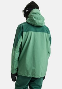 Groene skijas met een capuchon, met een matte textuur en een contrasterend donkerder groene schouderpaneel. Zwarte handschoenen en een muts gedragen.