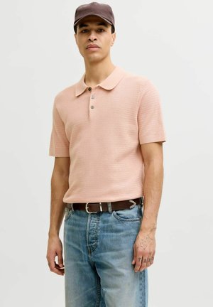 Jeune homme portant un polo manches courtes rose pâle, un jean bleu, une ceinture marron, une casquette marron foncé et des tatouages visibles sur les mains sur un fond clair.