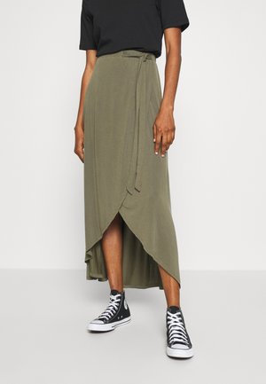 Osoba nosząca oliwkowozieloną spódnicę kopertową z asymetrycznym dołem oraz czarne wysokie trampki, w połączeniu z czarnym topem z krótkim rękawem.