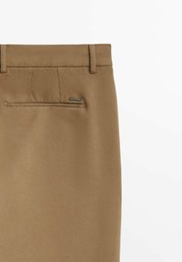 Zbliżenie na tył beżowych spodni z widocznym pasem, posiadającym szlufki na pasek oraz jedną kieszeń wpuszczaną z małą metalową metką marki.