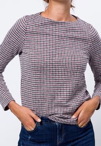 Persona que lleva una blusa de manga larga con estampado de pata de gallo en blanco, negro y burdeos, con jeans oscuros y las manos en los bolsillos.