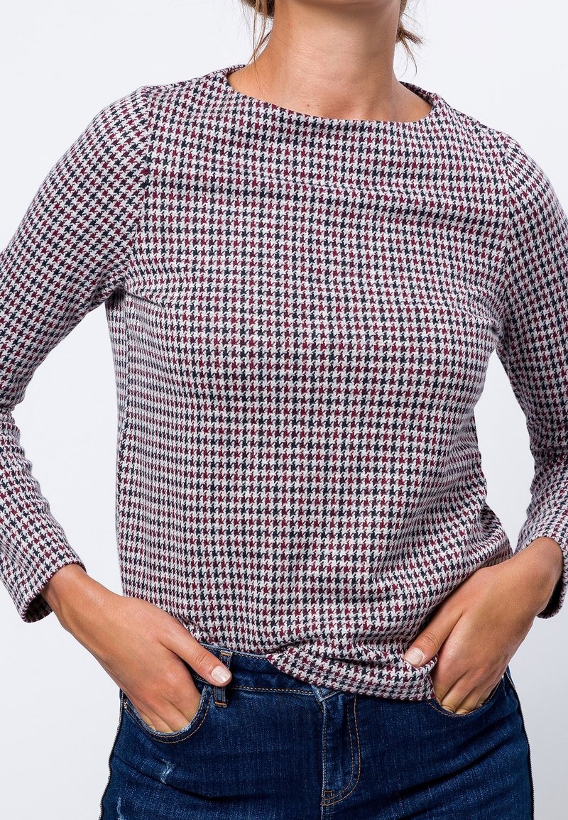 Persona que lleva una blusa de manga larga con estampado de pata de gallo en blanco, negro y burdeos, con jeans oscuros y las manos en los bolsillos.