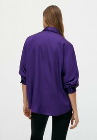 Lilla satinbluse med korte, op-rullede ærmer, klassisk krave og afslappet pasform, der har en glat tekstur og subtil glans.