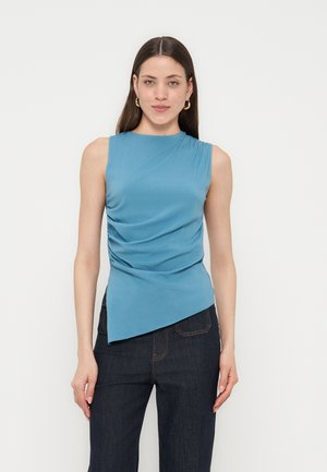 Γυναίκα με μακριά καστανά μαλλιά που φοράει χρυσούς κρίκους στα αυτιά, μπλε αμάνικη μπλούζα με ρίγες και ασύμμετρο τελείωμα και σκούρο μπλε τζιν.