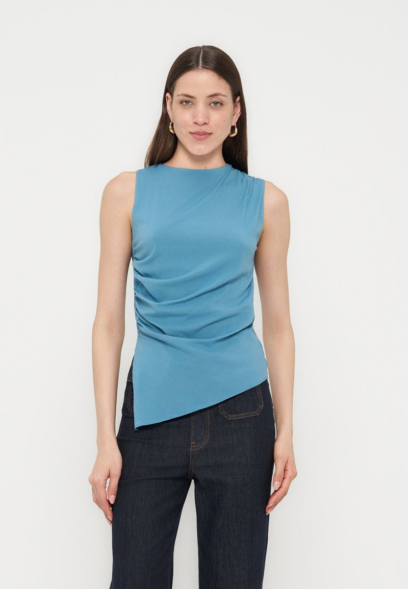 Γυναίκα με μακριά καστανά μαλλιά που φοράει χρυσούς κρίκους στα αυτιά, μπλε αμάνικη μπλούζα με ρίγες και ασύμμετρο τελείωμα και σκούρο μπλε τζιν.