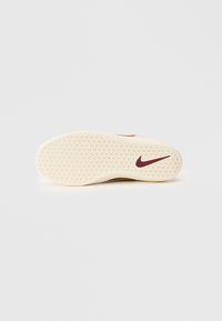 Hvit gummisol av en sneaker med bikake mønster og en maroon Nike swoosh-logo på en helt hvit bakgrunn.