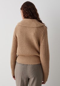 Kamelkleurige gebreide trui met een brede kraag, geribbelde zoom en een gestructureerd patroon, gecombineerd met gebloemde broek.