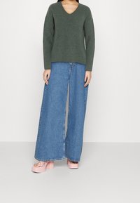 Maglione a coste di un verde scuro con scollatura a V; abbinato a jeans in denim leggeri e a gamba larga di un blu chiaro; sandali piattaforma rosa con motivo intrecciato.