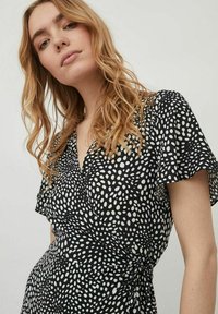 Rochie tip wrap, neagră cu buline albe, cu mâneci scurte și vaporoase, decolteu adânc în V și detaliu cu talie legată. Material ușor.
