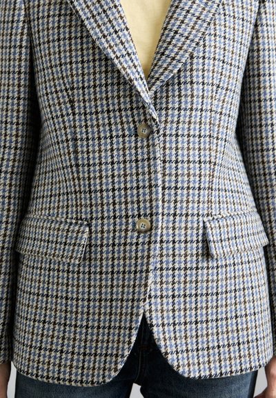 Ühe nööbiga bleiser, millel on sinine, pruun ja beež houndstooth muster, kinni nöödatud kahe nööbiga, klapi taskud, kantud beeži pluusi ja siniste teksade kohal.
