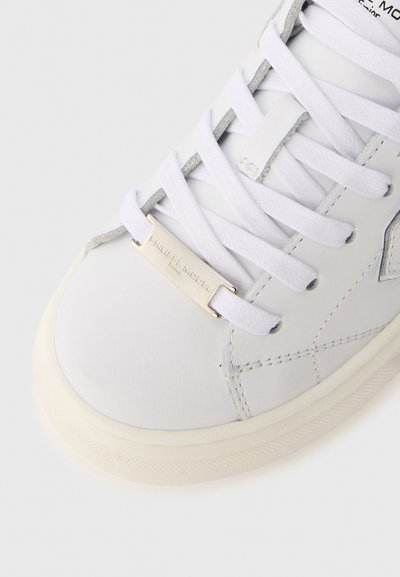 White leather sneaker with gray accents, featuring flat white laces, a metallic logo tag, and a textured rubber sole. Smooth finish.