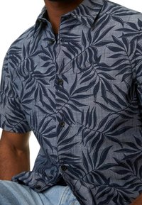 Mann in einem kurzärmeligen Hemd mit dunkelblauen Blattmustern auf einem hellblauen Hintergrund, kombiniert mit hellblauen Jeans.