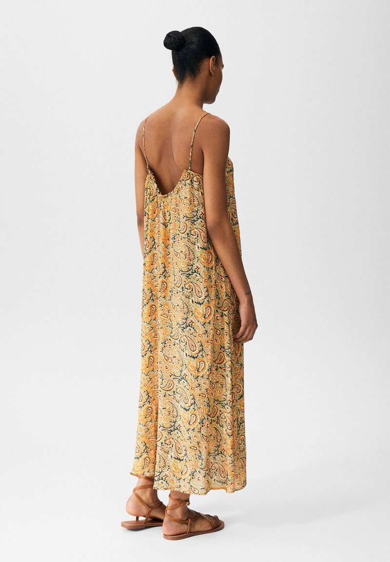 Robe maxi avec un motif cachemire en jaune, orange et vert. Dotée de fines bretelles et d'un dos nu. Associée à des sandales brunes simples.