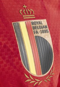 Maillot rouge avec un motif texturé, une couronne dorée et un logo avec des rayures noires, jaunes et rouges, et des accents de laurier.