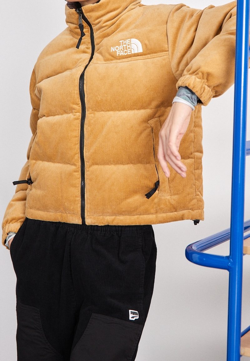 Person som bär en beige kordflors The North Face puffjacka och svarta Puma-byxor, lutad mot ett blått metallräcke.
