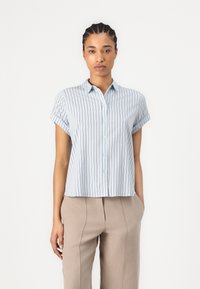 Camicia azzurra a maniche corte con righe bianche verticali, chiusura a bottoni e colletto. Indossata con pantaloni beige su misura.
